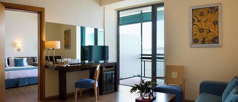 Habitación de hotel con cama, escritorio con televisor, balcón con vista y un cuadro en la pared.