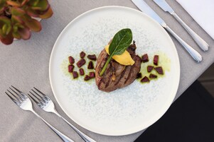 Un plato con un trozo de carne, una hoja y algunas guarniciones sobre un plato blanco texturizado.