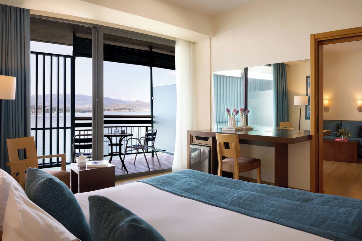 A hotel room with a bed, a desk, a chair, a small table, and a view of the outdoors.