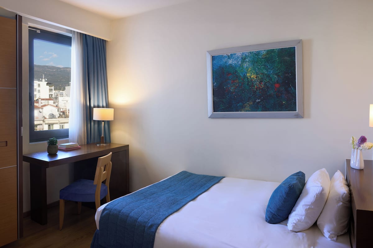 A hotel room with a bed, a desk, a chair, a window with a view of buildings, and a painting on the wall.