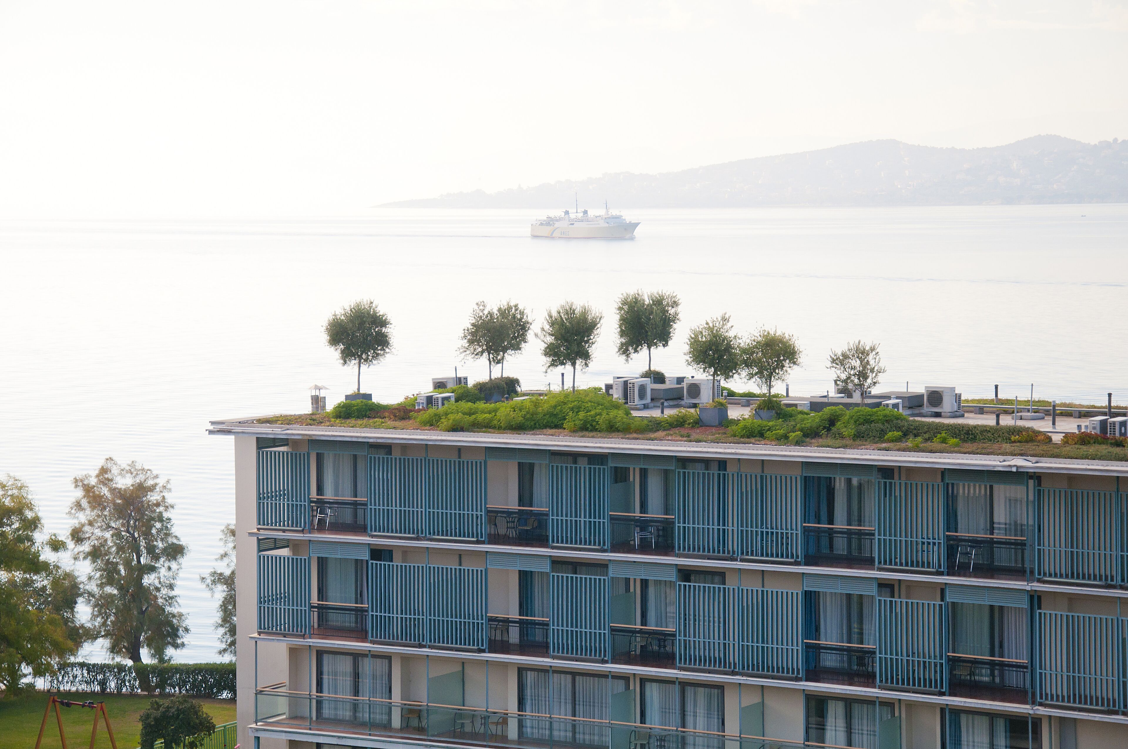 A multi-story building with balconies, a rooftop garden with trees, and a view of a body of water with a ship in the distance.