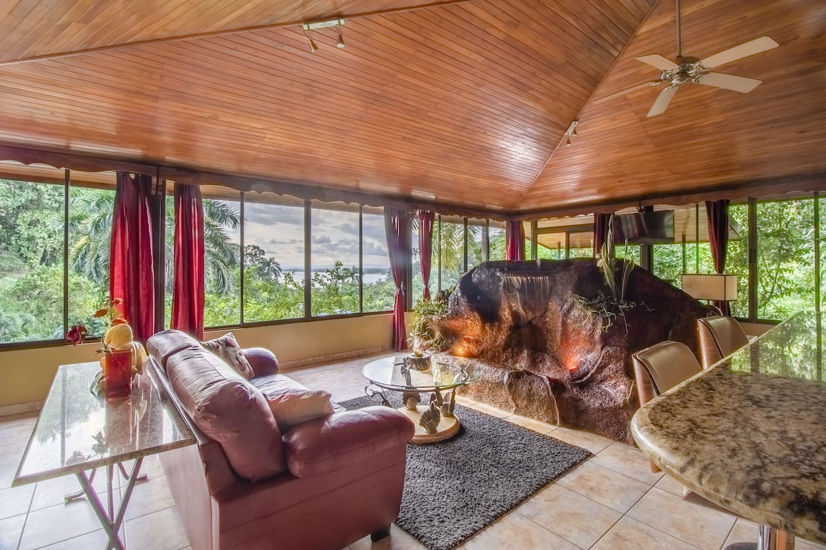 Une pièce spacieuse avec un plafond en bois, une grande fenêtre avec des rideaux, un canapé, une table basse en verre, une table à manger et une cheminée.