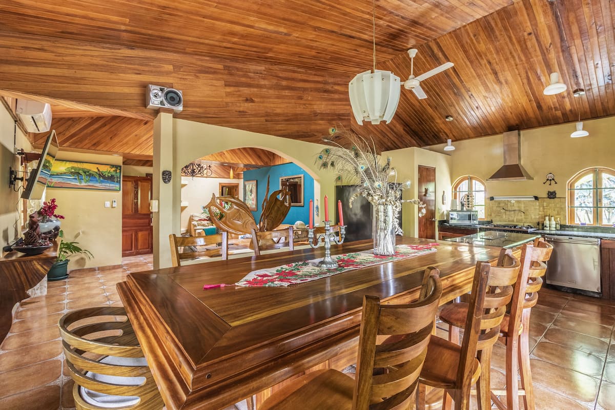 Un grand espace à manger avec une table et des chaises en bois, un ventilateur de plafond et une cuisine en arrière-plan.
