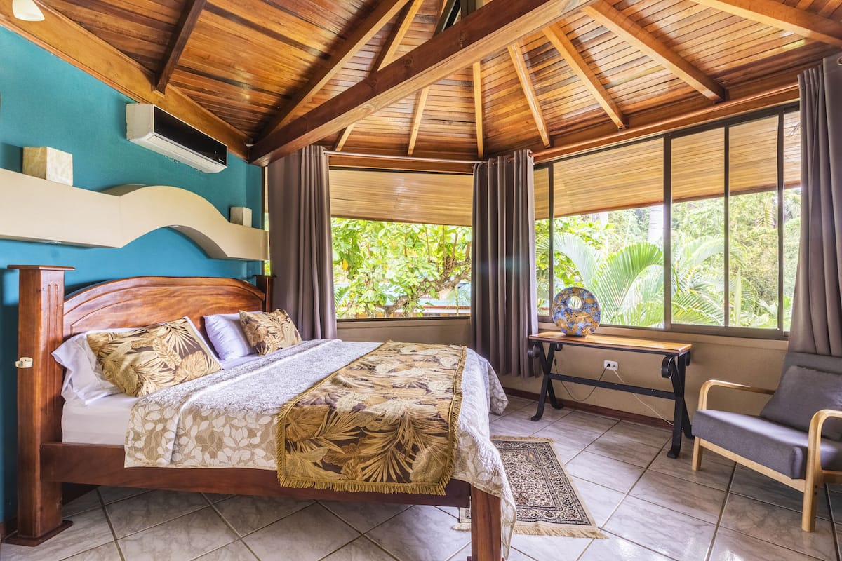 Une chambre avec un plafond en bois, un lit recouvert d’une literie à motifs, une fenêtre avec des rideaux et une vue sur la verdure à l’extérieur.