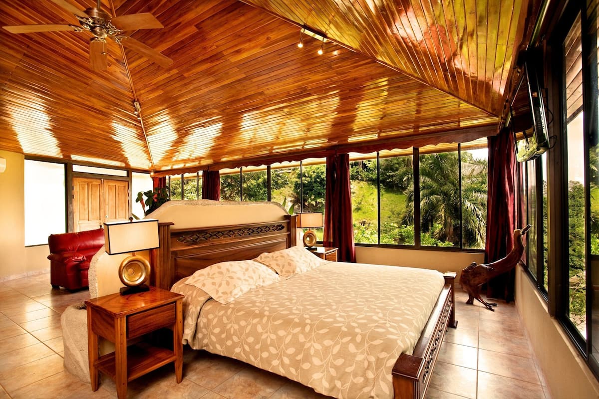 Une chambre avec un grand lit, un plafond en bois, une table de chevet, une lampe, un fauteuil rouge et une vue sur la verdure à l’extérieur.