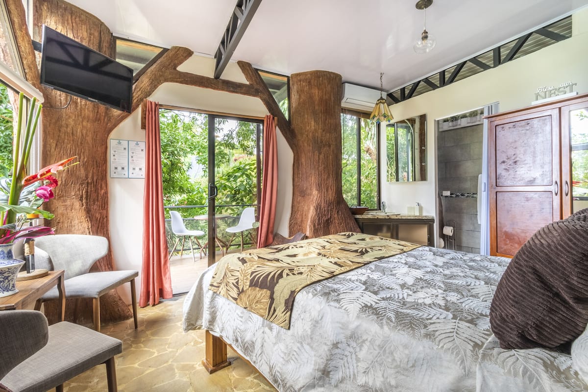 Une chambre avec un grand lit, un téléviseur fixé sur le tronc d’un arbre, un bureau avec une chaise et une vue sur l’extérieur.