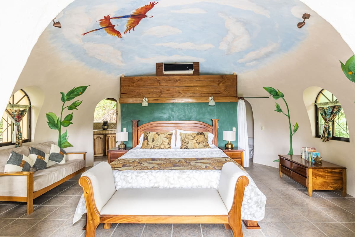 Une chambre spacieuse avec un grand lit, une tête de lit en bois et une fresque représentant des oiseaux au plafond.