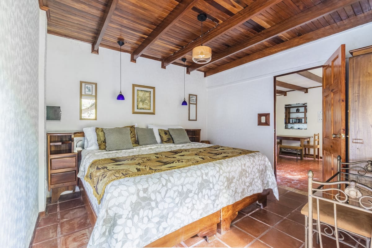 Une chambre avec un plafond en bois, un lit recouvert d’une couvre-lit à motifs floraux et un coin repas visible à travers une porte ouverte.