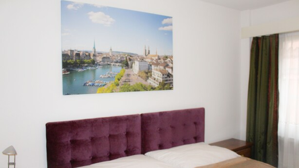 A hotel room with a large bed, a headboard, a nightstand, a lamp, and a cityscape painting on the wall. - Hotel Villette (Zürich)