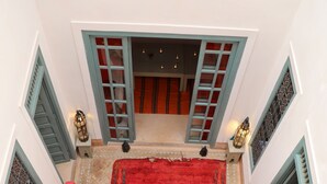 Un balcón espacioso con alfombra roja, dos sofás rojos, una mesa blanca con un florero y vista al interior a través de puertas abiertas.