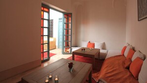Habitación de hotel con cama, una mesa de madera con velas, una mesita con un florero y vistas a través de una puerta abierta.