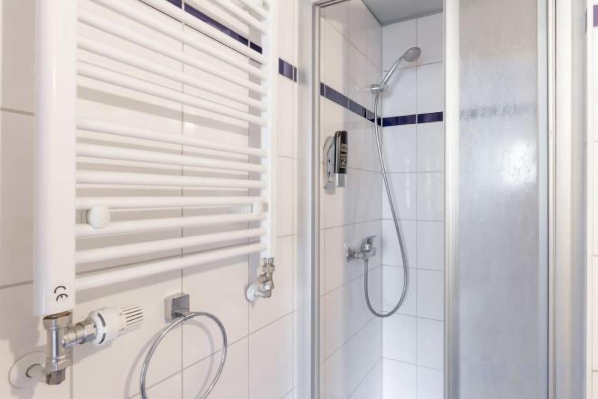 Un baño con cabina de ducha de vidrio, un radiador blanco y una tubería visible.