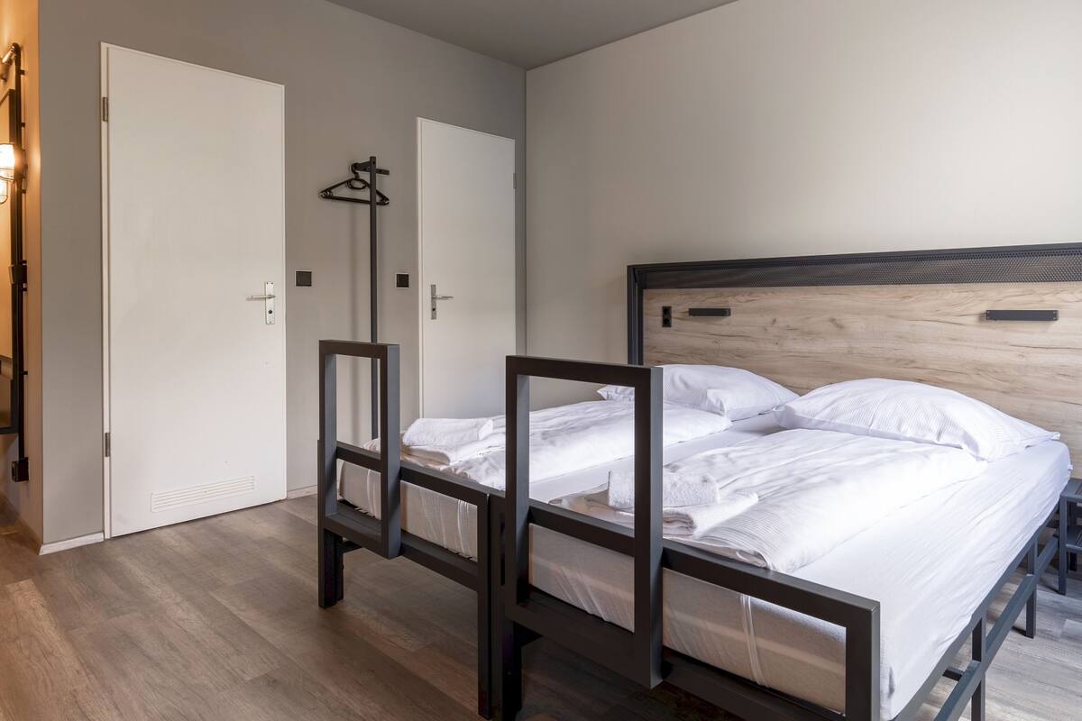 Habitación de hotel con estructura de cama de madera, ropa de cama blanca, una puerta con perchero y suelo de tonos claros.