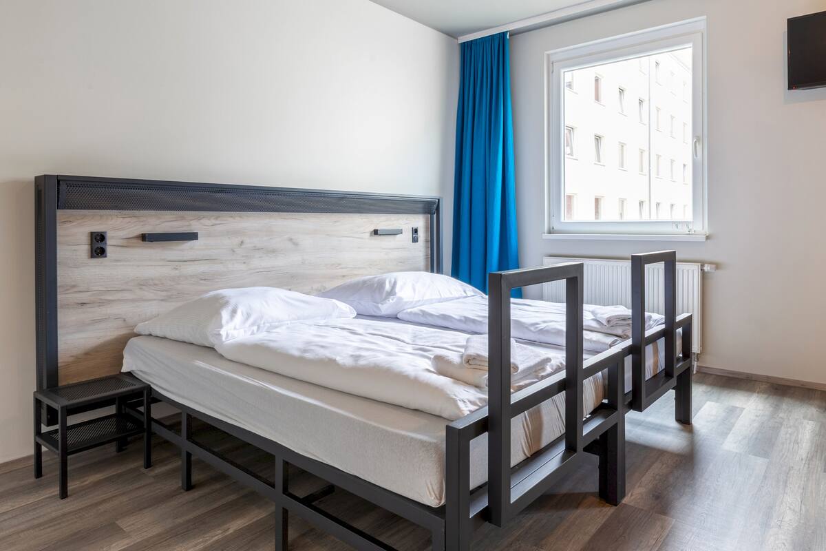Una cama individual con cabecera de madera, una mesita de noche pequeña y una ventana con cortinas azules.