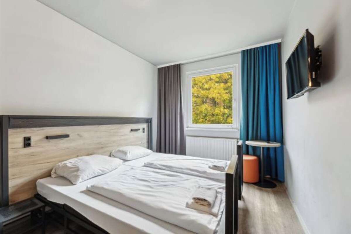 Una habitación de hotel con una cama, un televisor montado en la pared, una ventana con cortinas y una mesita de noche con lámpara.