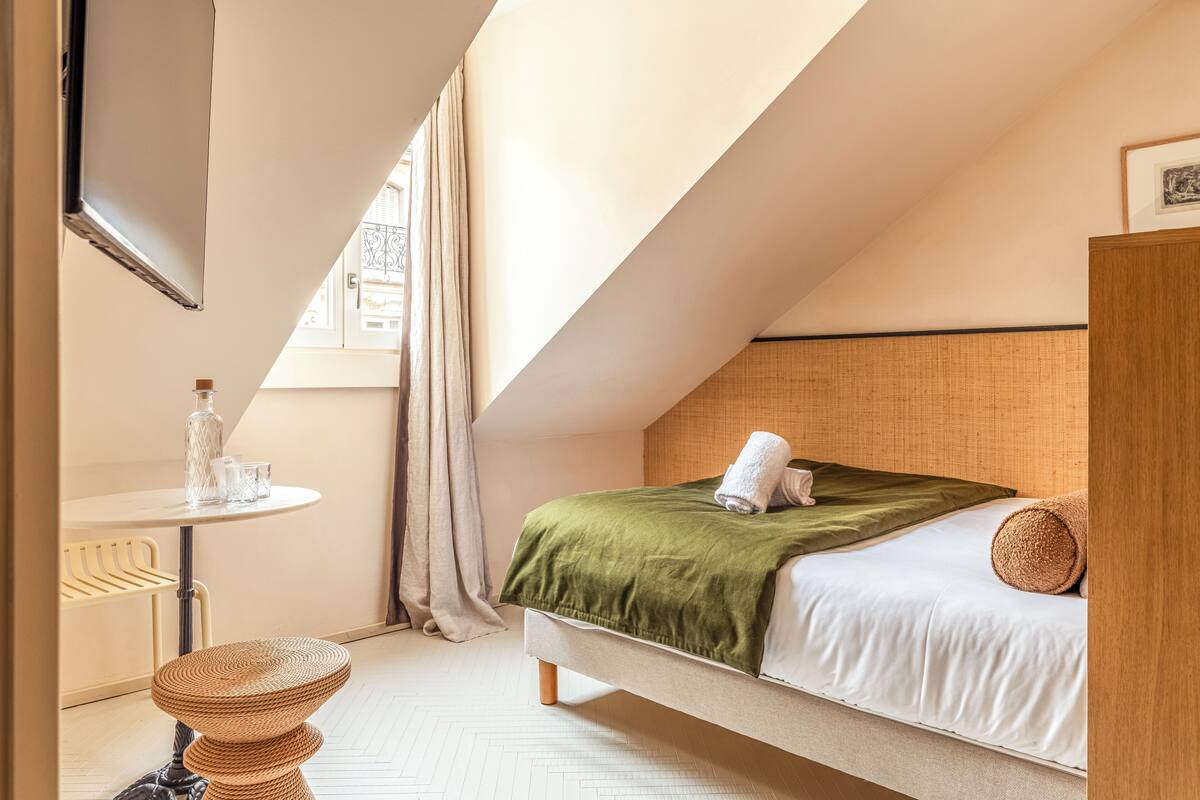 Ein Schlafzimmer mit einem Bett, einem kleinen Tisch mit einer Flasche, einem Sessel und einem Fenster mit Vorhängen.