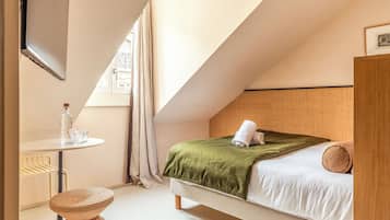 Ein Schlafzimmer mit einem Bett, einem kleinen Tisch mit einer Flasche, einem Sessel und einem Fenster mit Vorhängen.