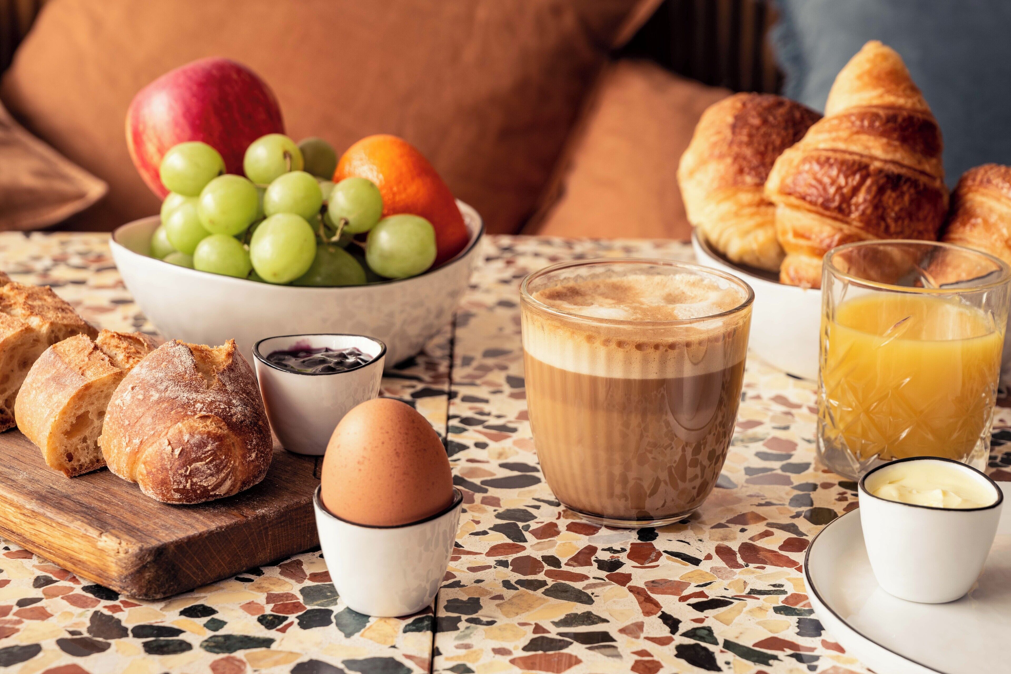 Een ontbijttafel met brood, croissants, fruit, koffie en sap op een betegelde tafel.