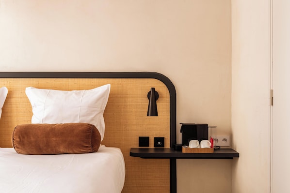 Quarto de hotel com cama, almofadas, mesa de cabeceira com abajur, um pequeno alto-falante e cafeteira.