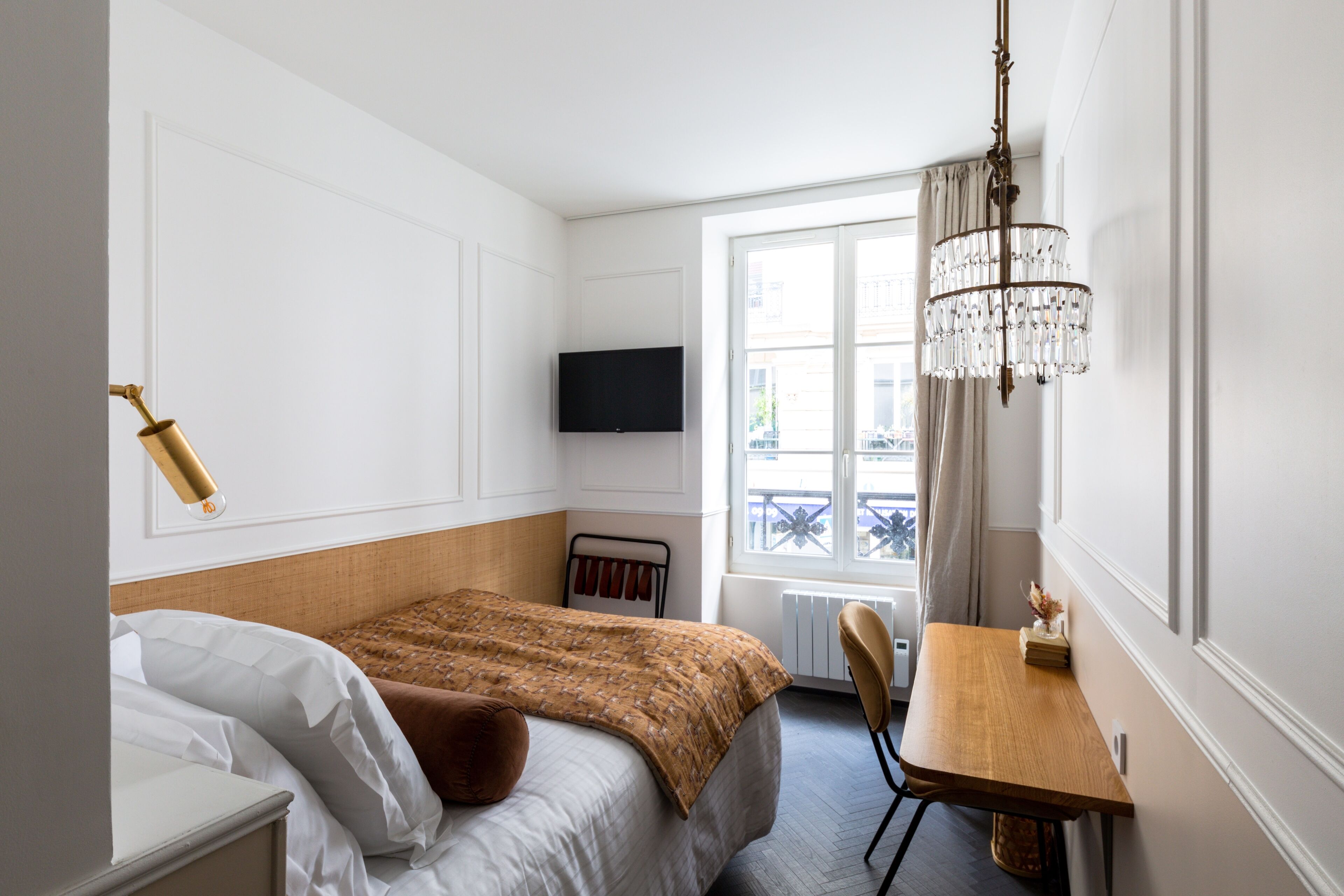Een hotelkamer met een bed, een bureau, een kroonluchter, een televisie en een raam met gordijnen.