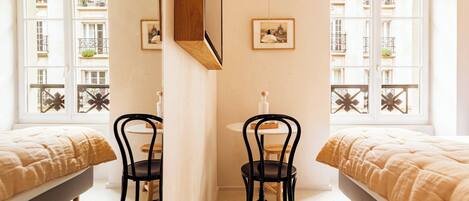 A room with two beds, a wooden chair, a small table, and a window with a view of balconies.