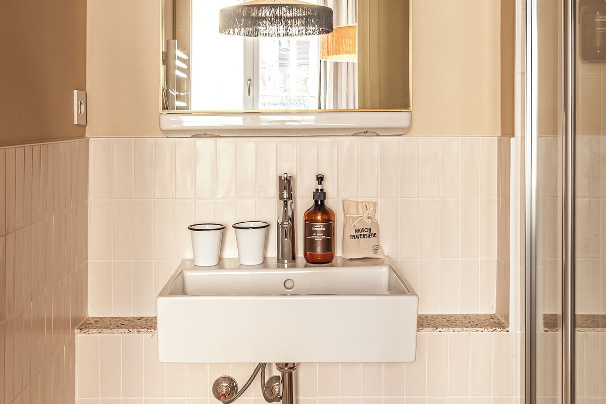 Une salle de bain avec un lavabo blanc, un miroir surmonté d’une arche incurvée et un espace douche.