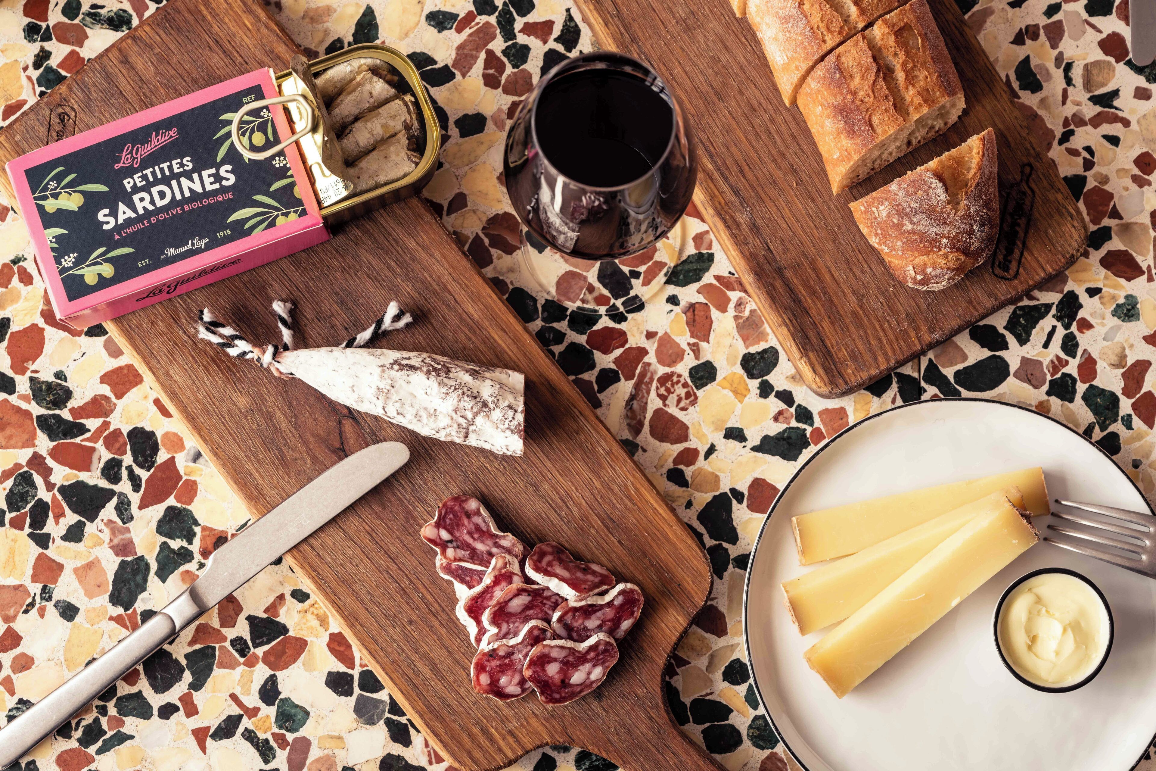 Una tabla de madera con una lata de Pequeñas Sardinas, un tarro de aceitunas, una hogaza de pan y una copa de vino tinto.