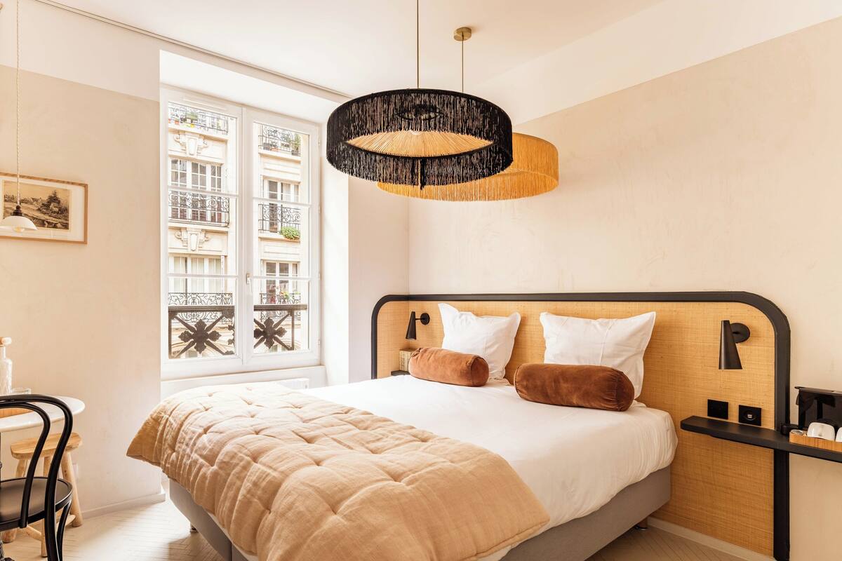 Ein Schlafzimmer mit einem großen Bett, einem Nachttisch, einer Hängeleuchte und einem Fenster mit Blick auf Gebäude.