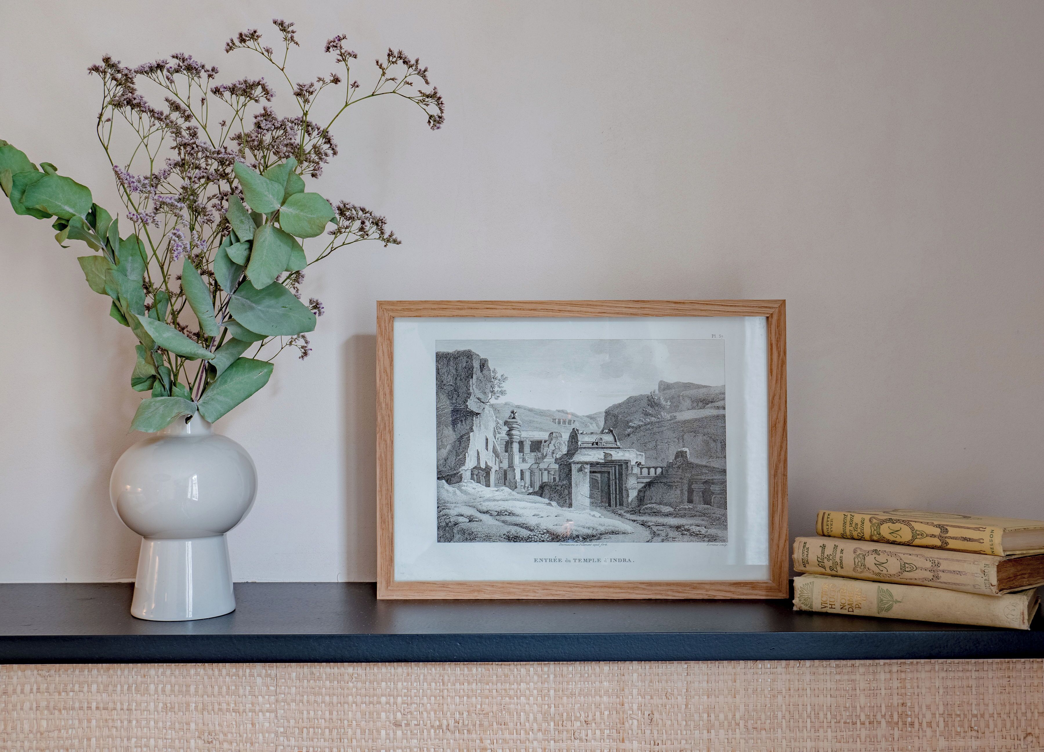 A framed black and white photograph of an ancient temple, a white vase with green leaves, and a stack of old books on a shelf.