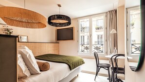 Un dormitorio moderno con cama, televisor, zona de comedor con sillas y ventanas amplias con vista a edificios.