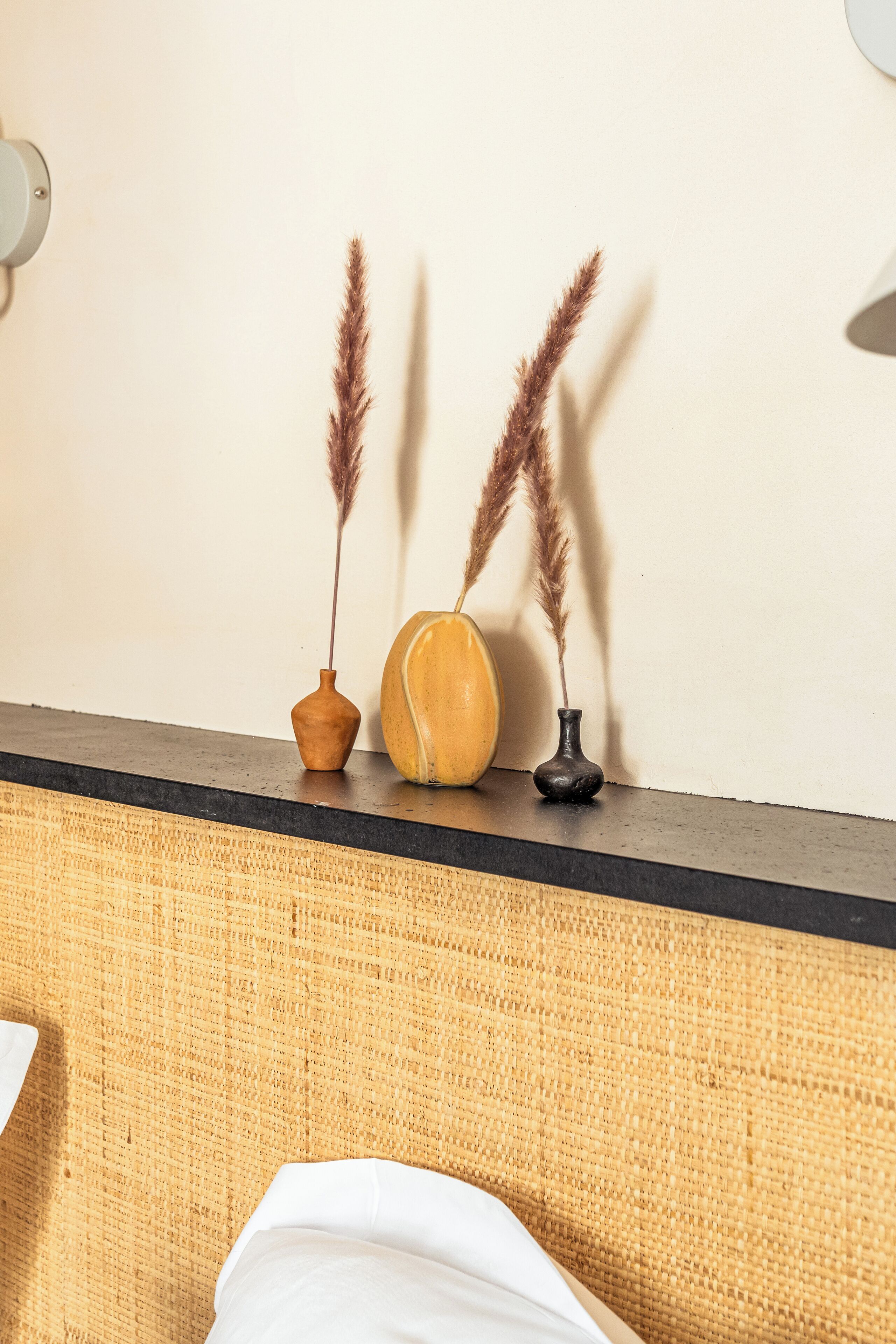 A bed with a woven headboard, two vases with pampas grass, and a wall-mounted light fixture.