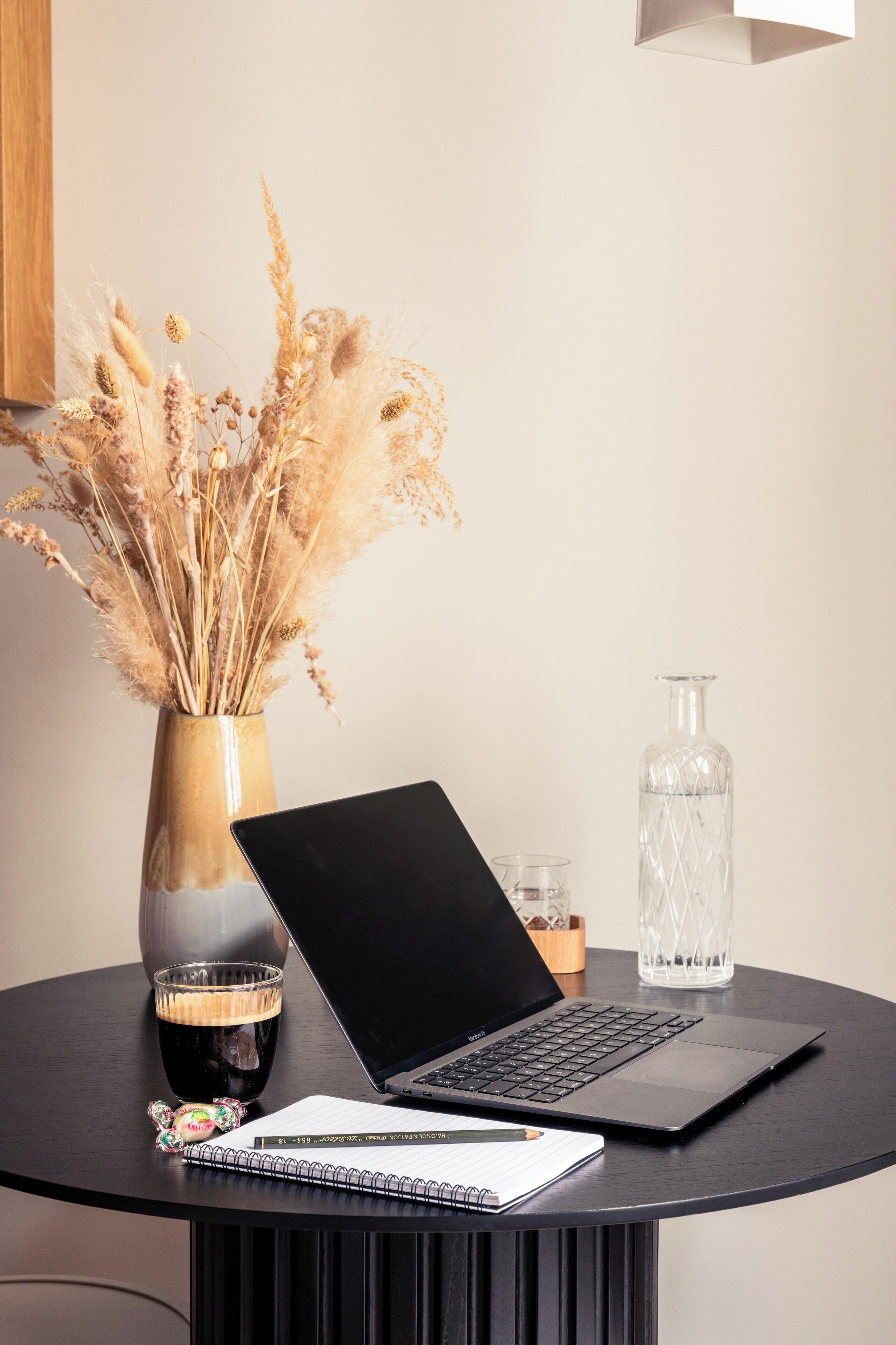 A laptop on a round table with a notebook, a glass of dark liquid, a decorative vase with dried plants, and a clear bottle.