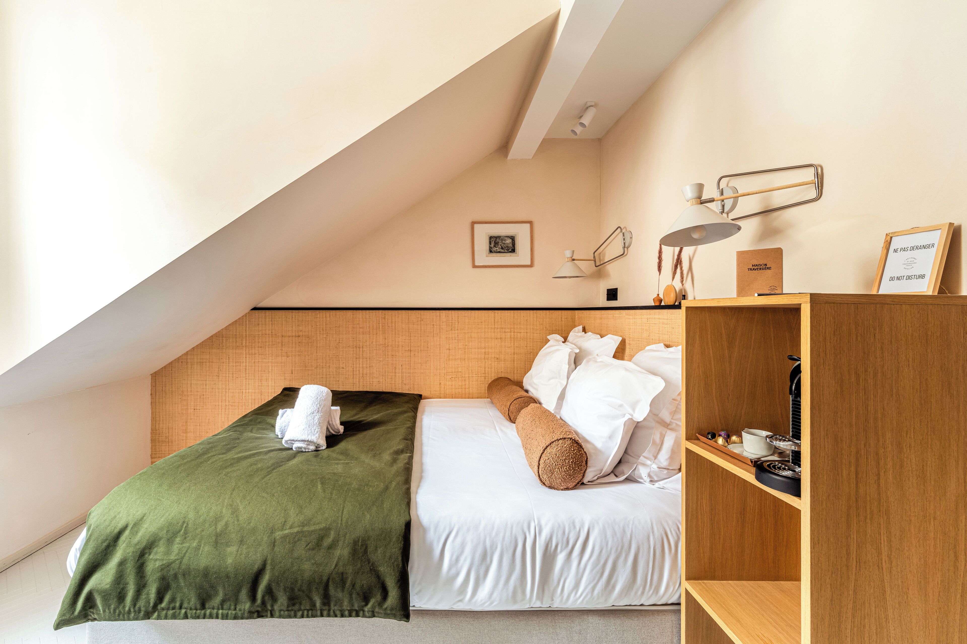 Una habitación de hotel con una cama, una manta verde, una mesita de noche de madera con una lámpara y un cartel enmarcado en la pared.