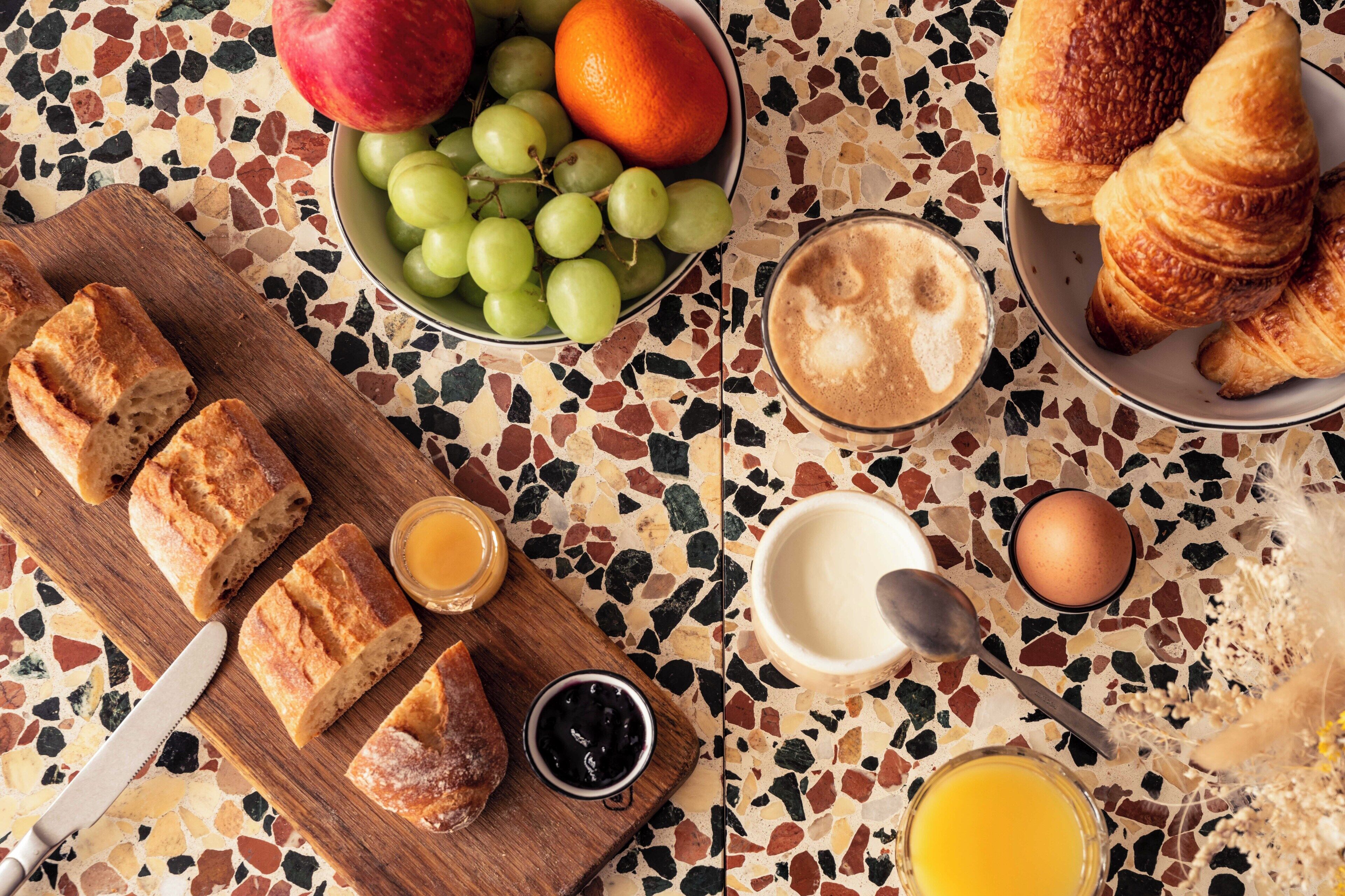 A breakfast setting with bread, fruit, croissants, coffee, and a glass of orange juice on a mosaic table.