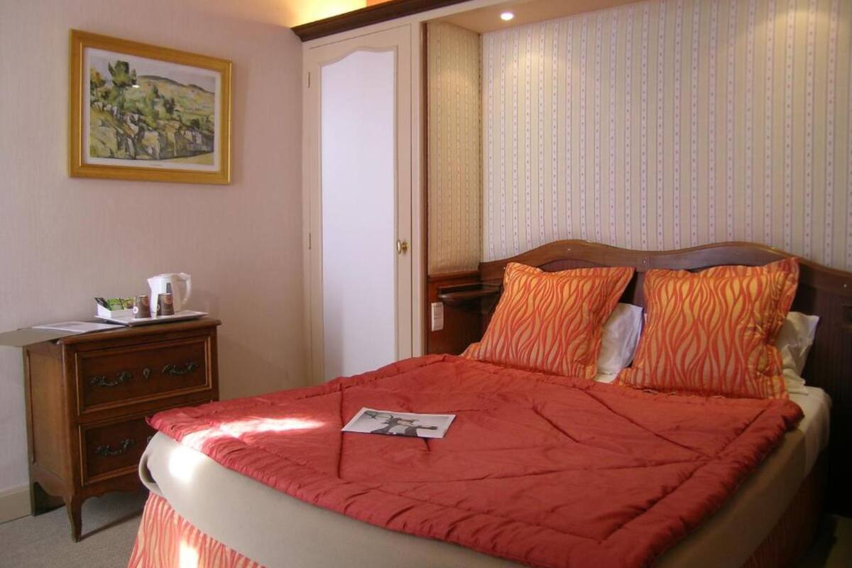 Een net opgemaakt bed met een rode deken, een houten nachtkastje met een koffiezetapparaat en een ingelijste foto aan de muur.