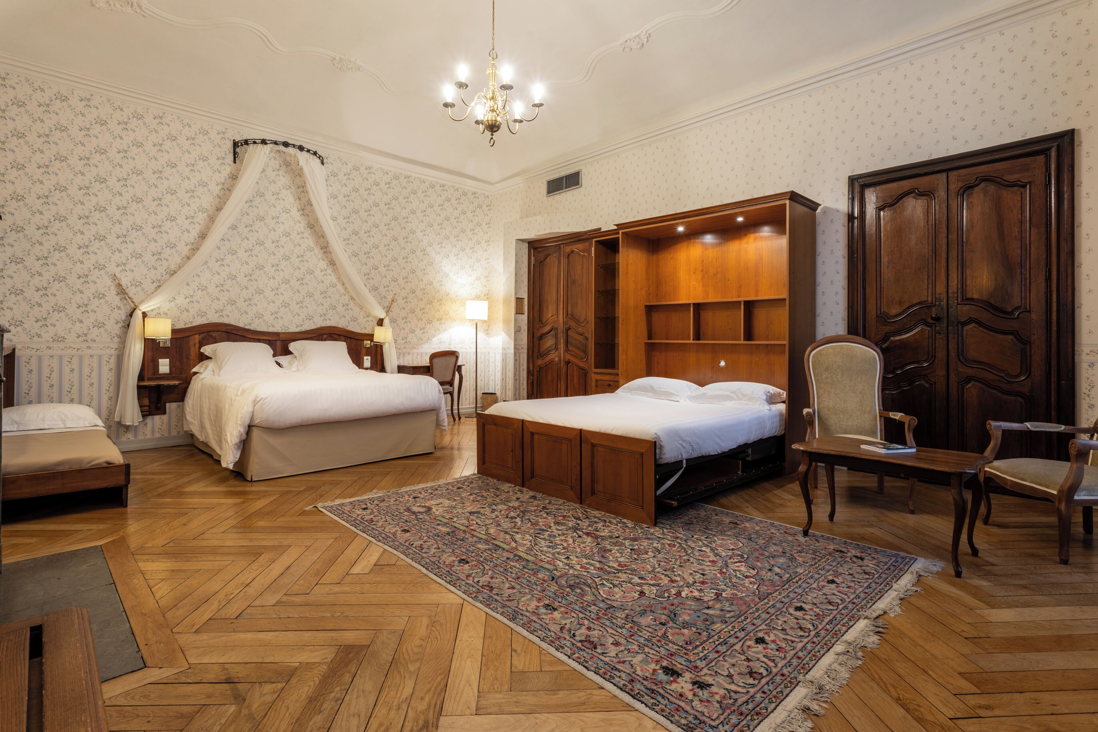 A hotel room with a large bed, a wooden wardrobe, a chair, a desk, and a patterned rug.