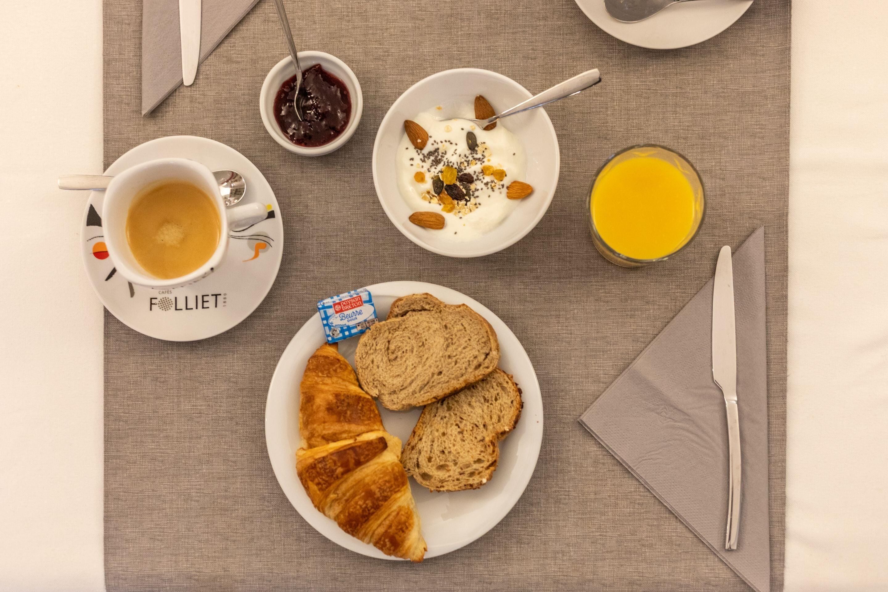 Un desayuno con croissant, pan, café, yogur con almendras y semillas, mermelada y jugo de naranja.