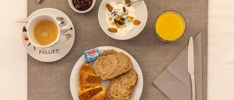 Un desayuno con croissant, pan, café, yogur con almendras y semillas, mermelada y jugo de naranja.