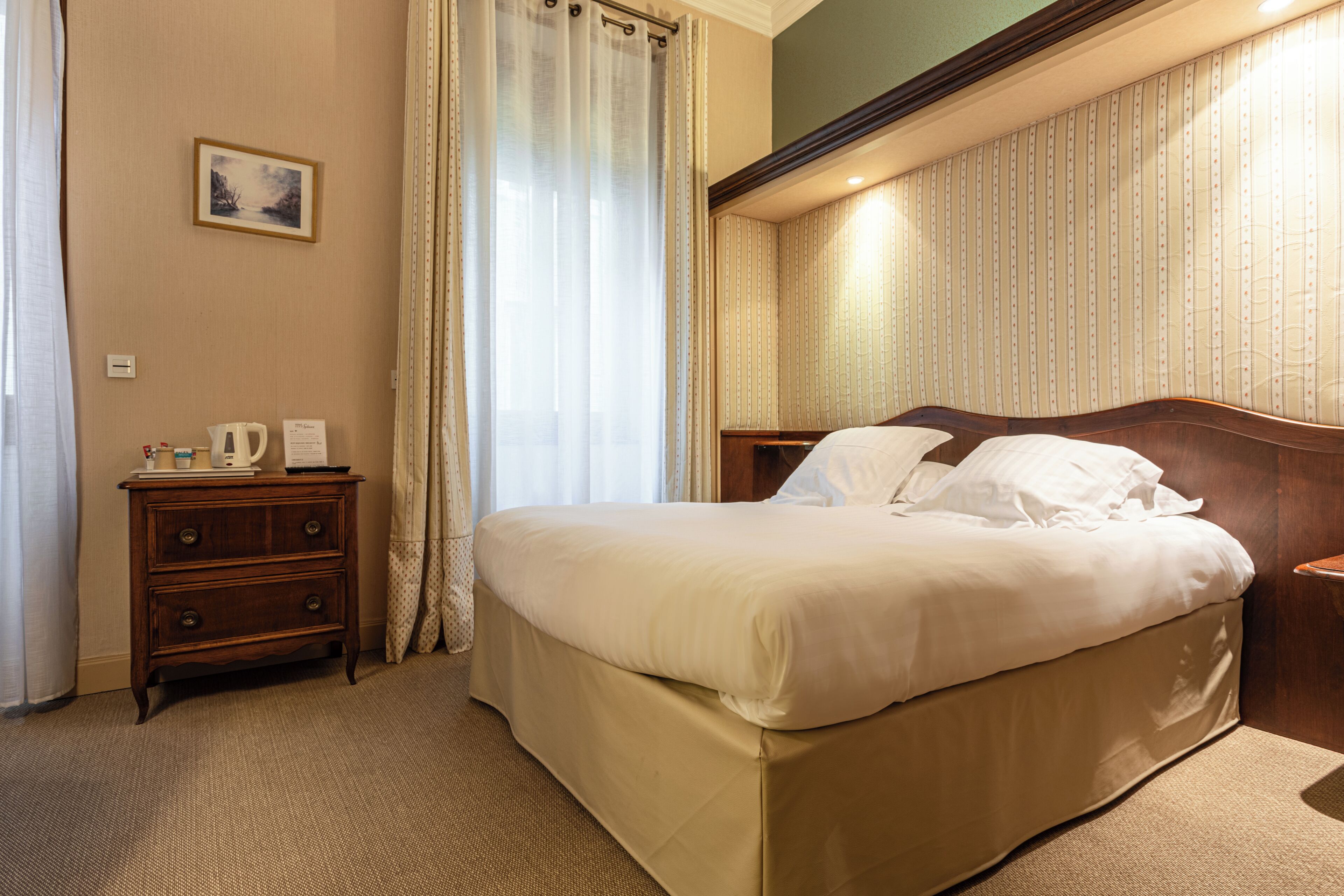 Habitación de hotel con una cama, una mesita de noche, un aparador de madera, un cuadro en la pared y una ventana con cortinas.