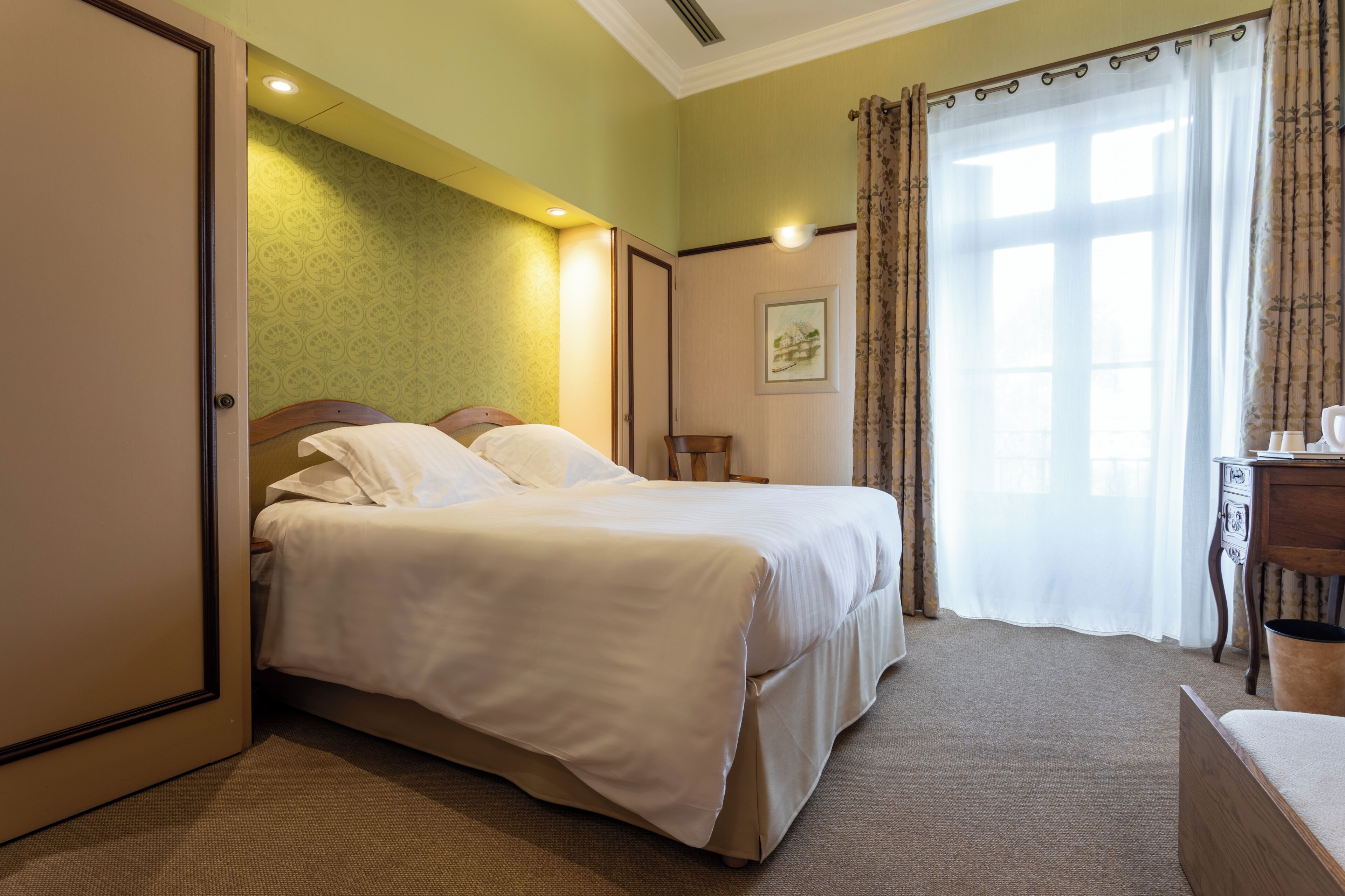 A hotel room with a bed, a desk, a chair, a picture on the wall, and a window with curtains.