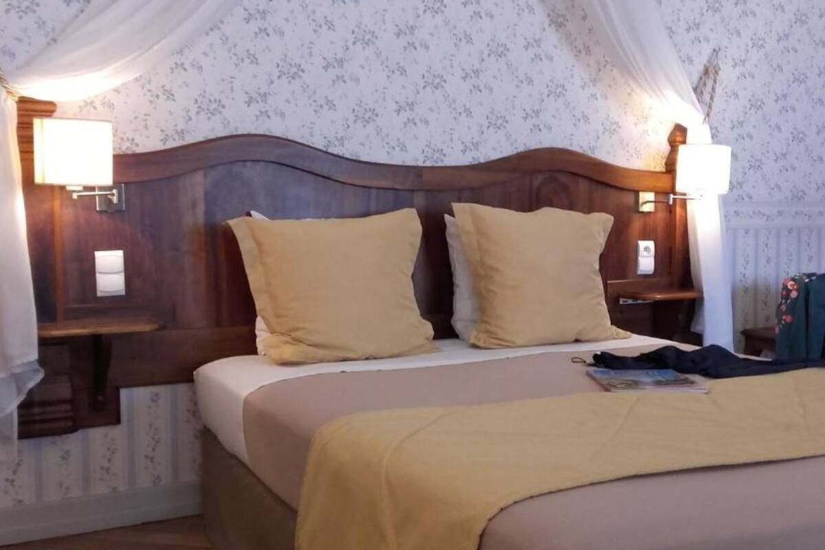 Een hotelkamer met een houten hoofdbord, twee bedlampen, een bed met een beige sprei en een nachtkastje met een tas en een boek.