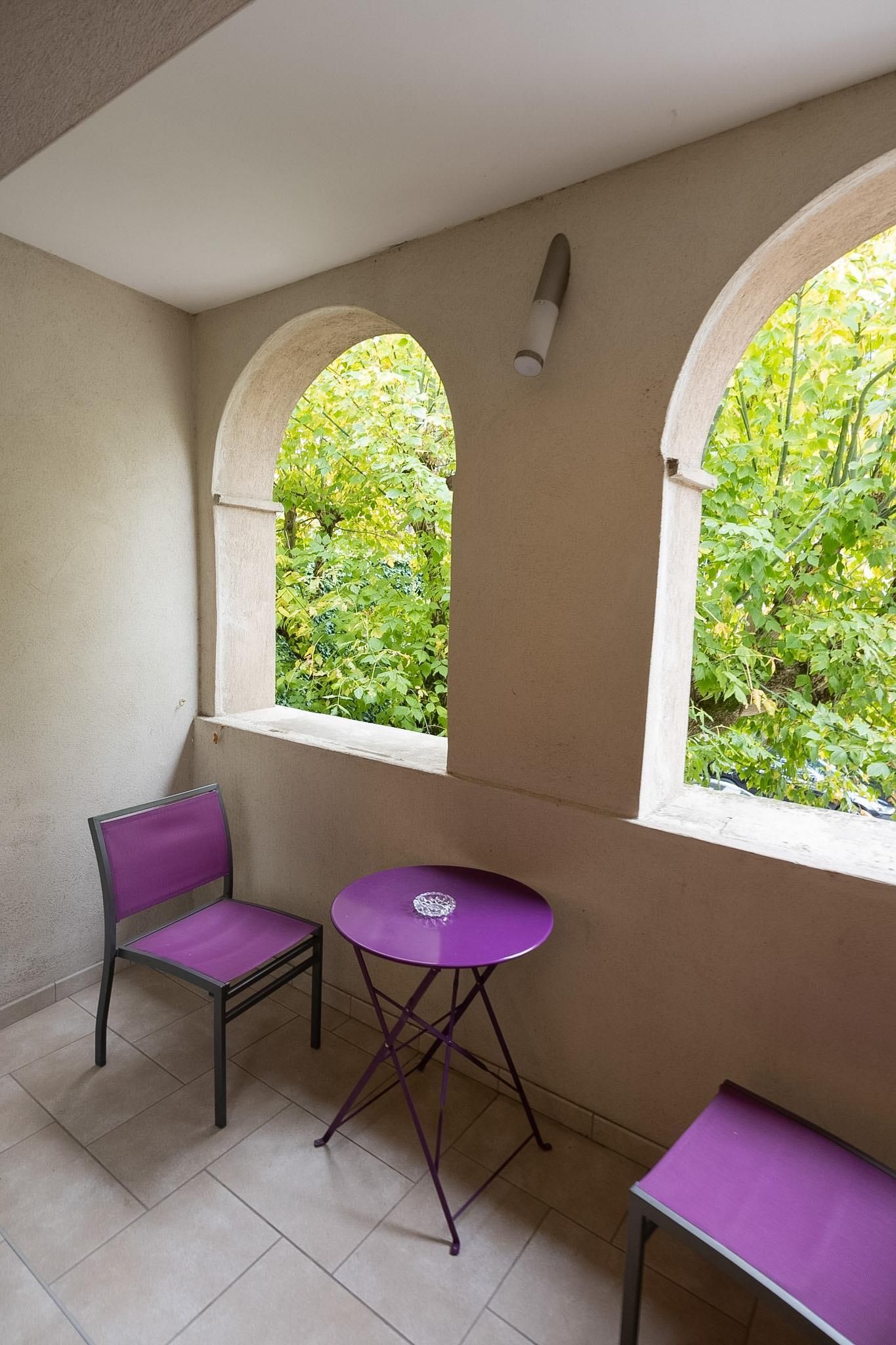 Un pequeño balcón con dos sillas moradas y una mesa, una ventana con vistas a zonas verdes, y una lámpara fijada a la pared.