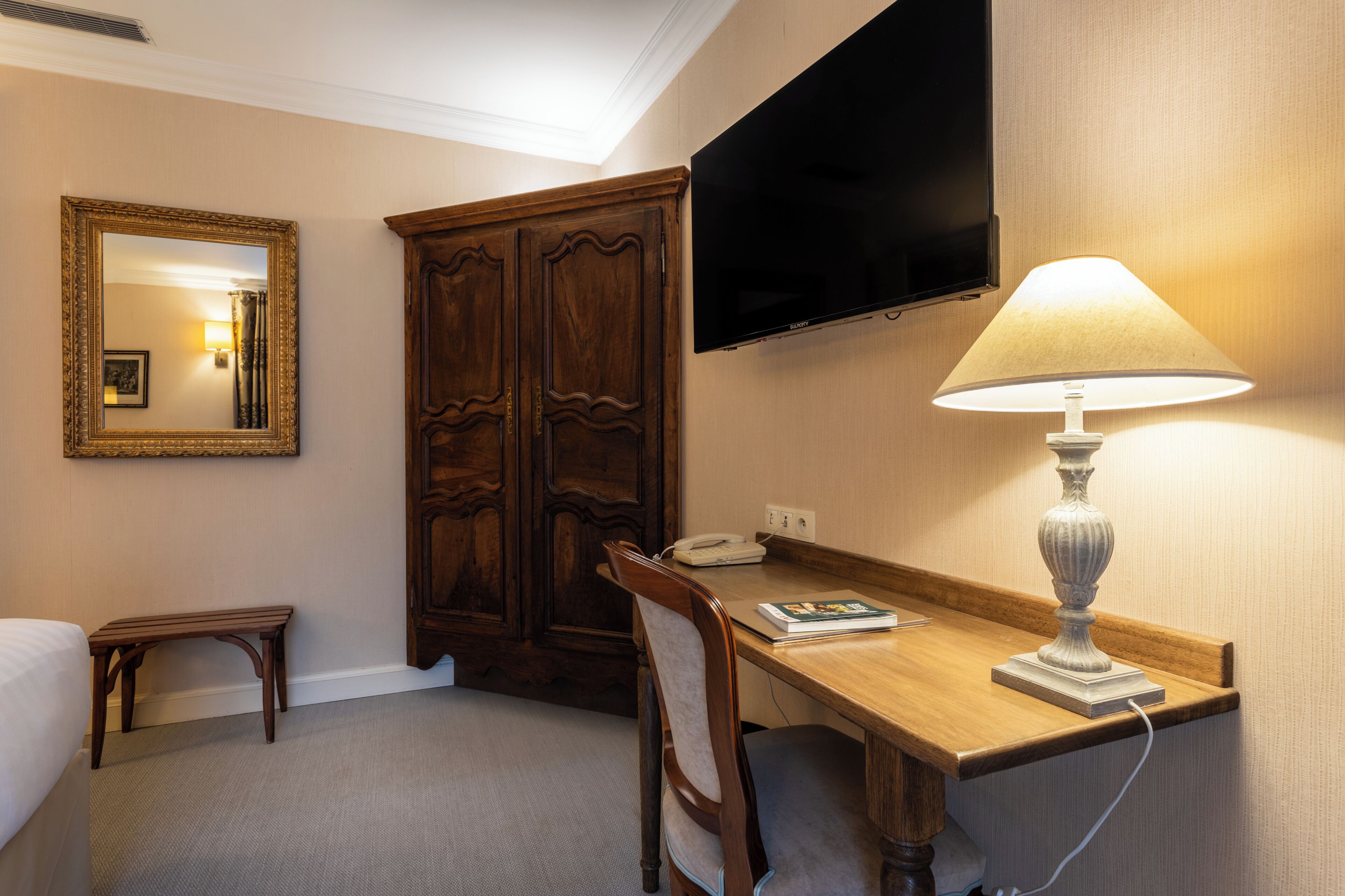 Habitación de hotel con escritorio de madera, una silla, una mesita de noche, una lámpara, un televisor de pantalla plana montado en la pared, un espejo grande y una pequeña mesita de noche con un teléfono y un libro.