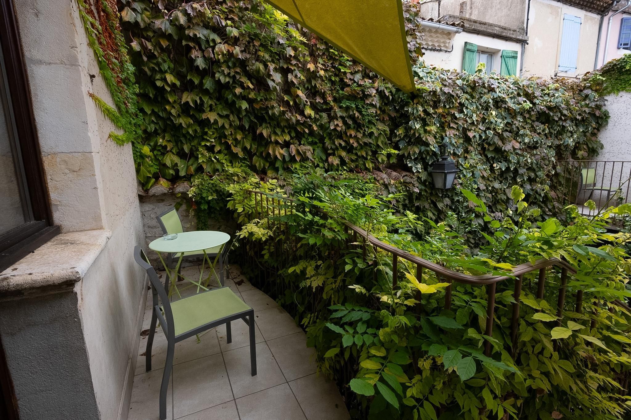 A small outdoor seating area with a table and chairs, surrounded by dense foliage and a yellow awning.