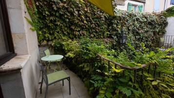 A small outdoor seating area with a table and chairs, surrounded by dense foliage and a yellow awning.