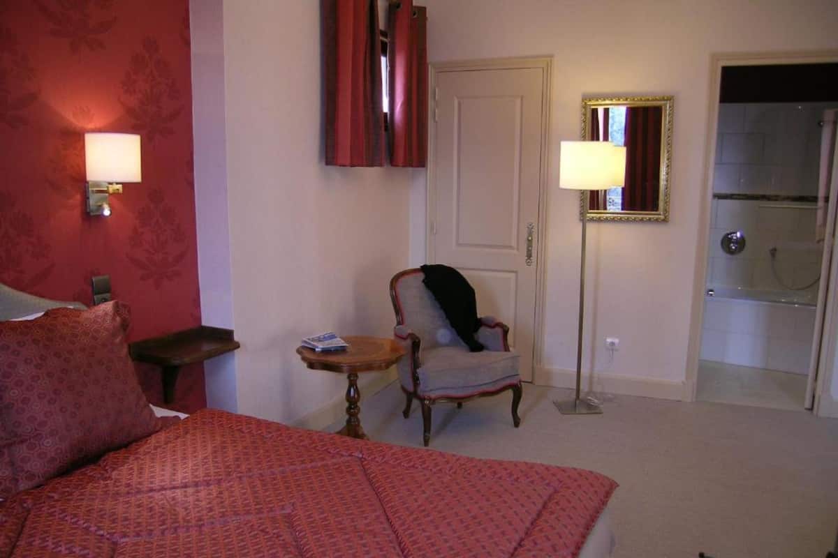 Een hotelkamer met een bed, een stoel, een klein tafeltje, een lamp, een spiegel en een badkamers deur.