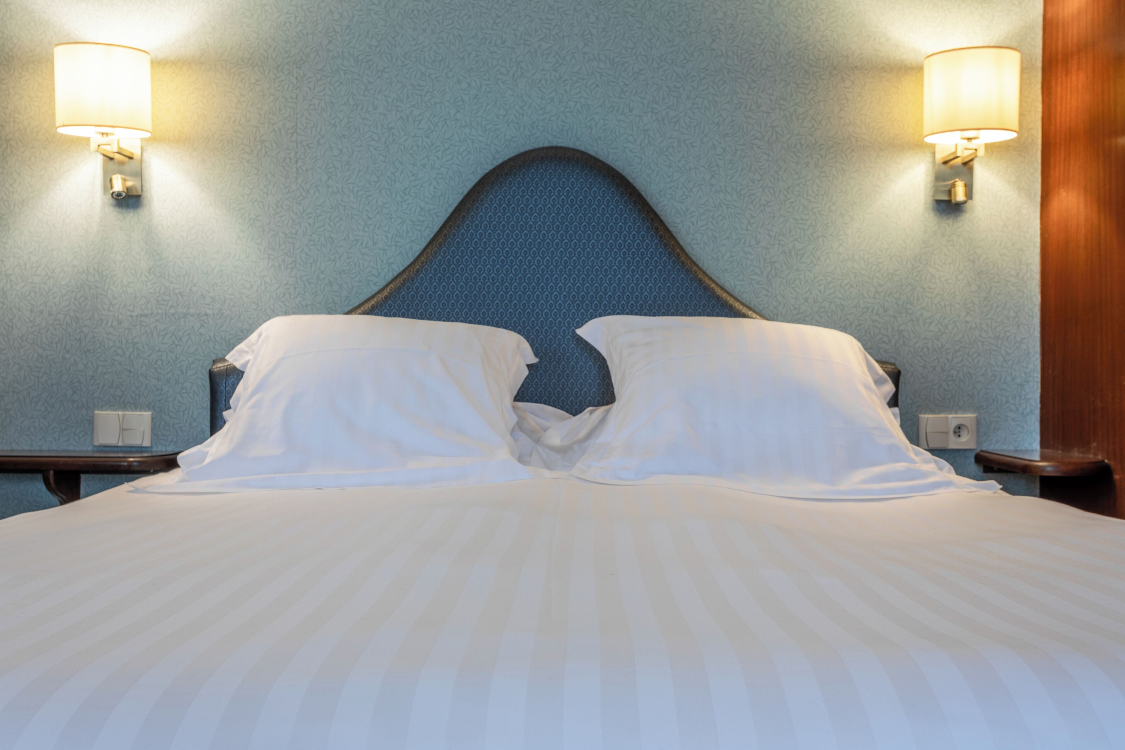 Une chambre d’hôtel avec un grand lit, deux lampes fixées au mur et un mur bleu à la texture particulière.