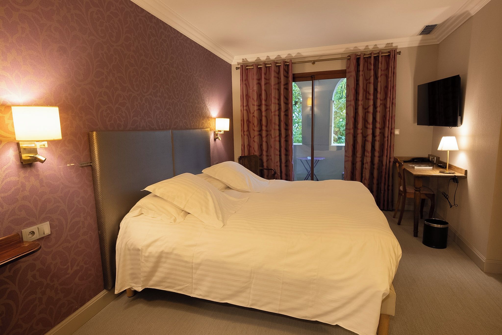A hotel room with a bed, a desk with a lamp, a television, and a view of greenery outside.