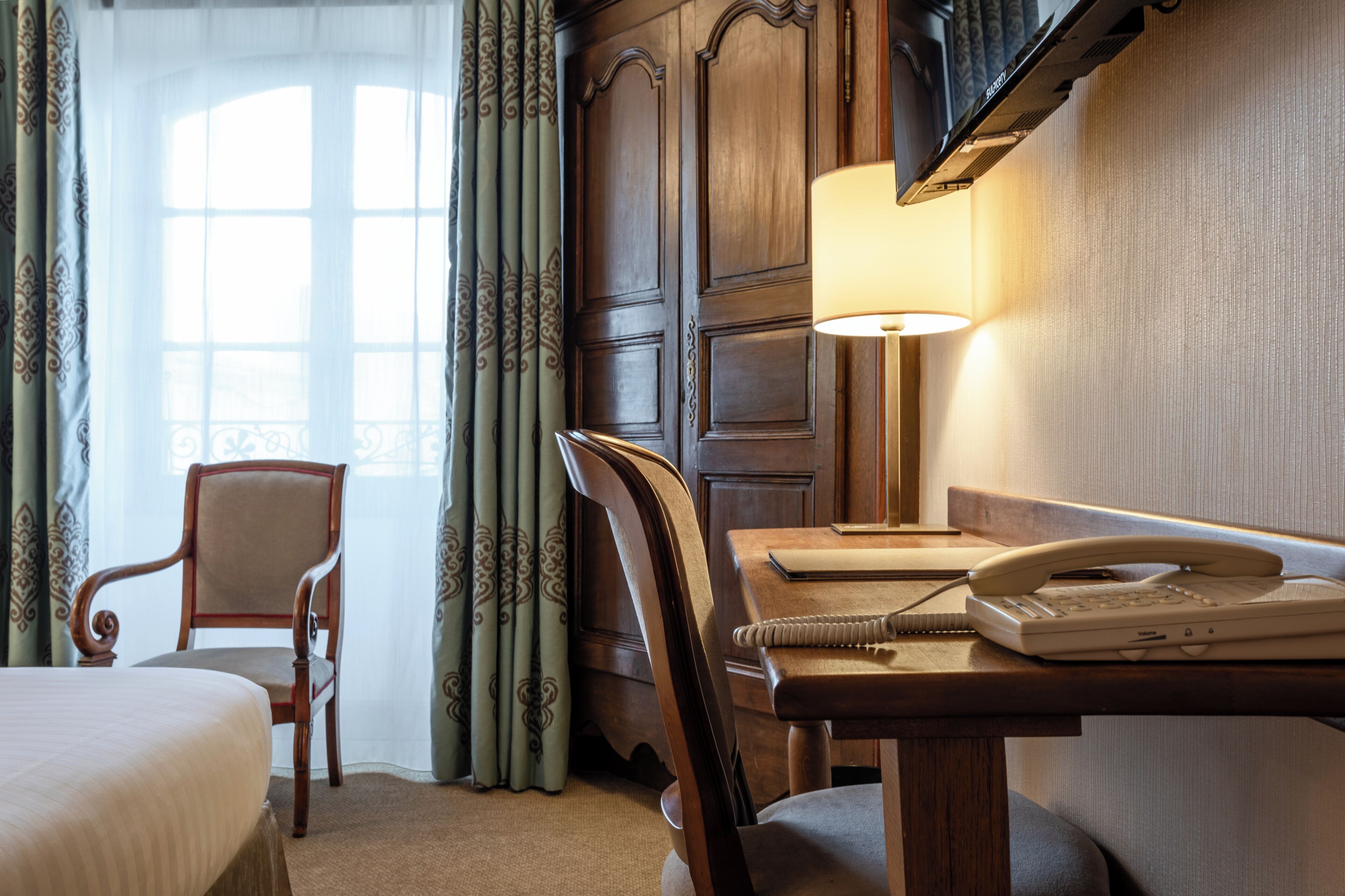 A hotel room with a bed, a desk with a lamp, a telephone, and a chair.