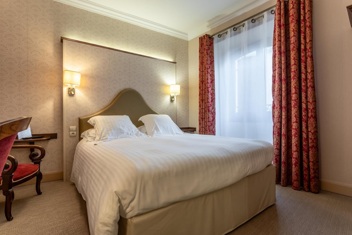 A hotel room with a large bed, two bedside lamps, a wooden chair, and a window with red curtains.